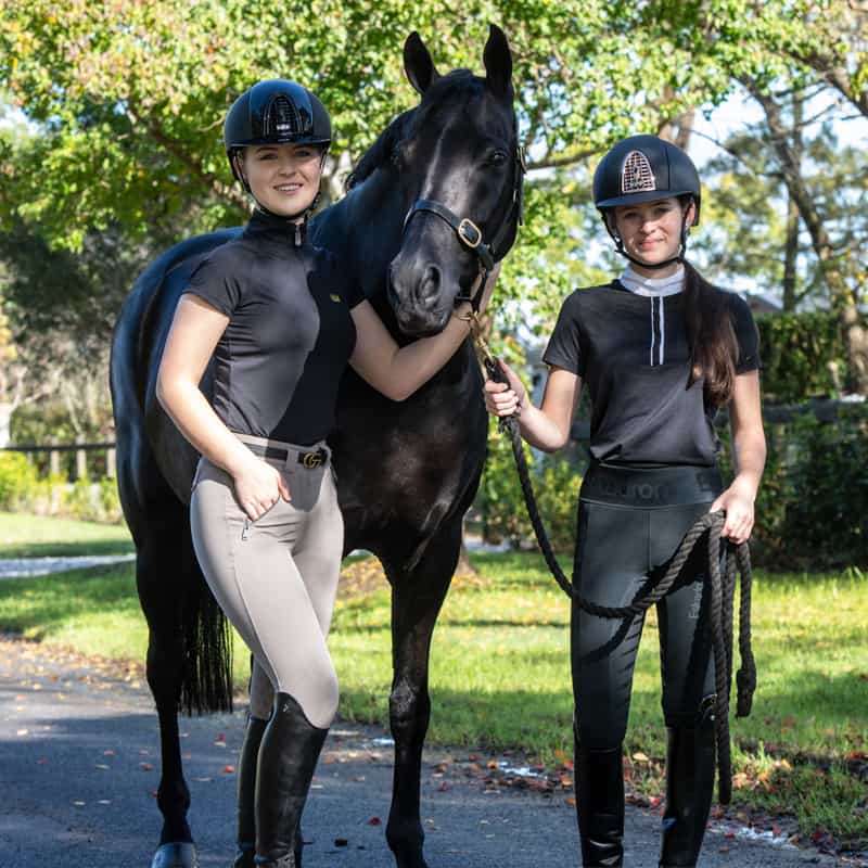 Image of two young women wearing equestrian gear available to purchase from Saddleworld Dural. They are standing with a chestnut horse between them.