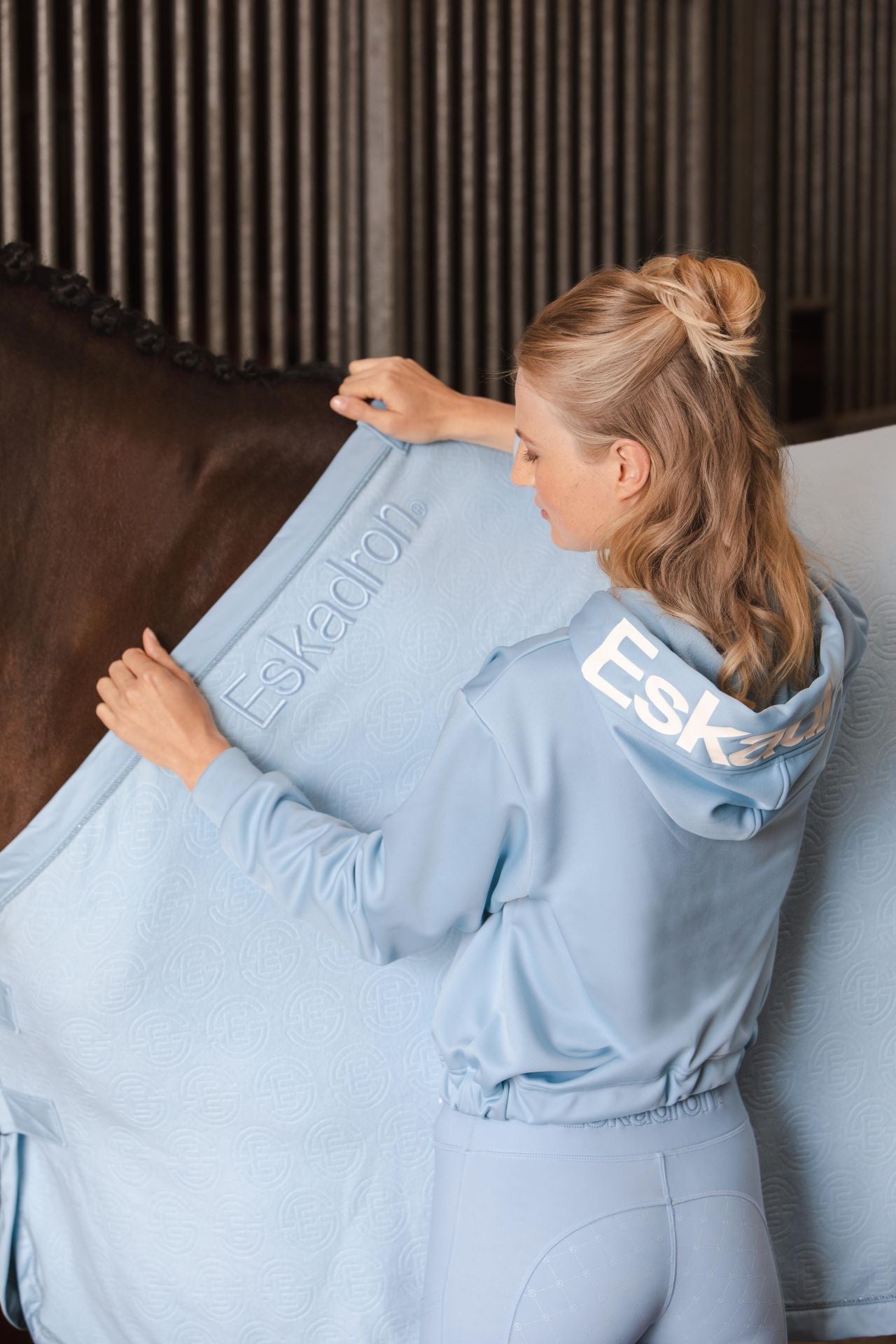 Image of a woman wearing the light blue hoodie from the Eskadron Reflexx 2023 collection, standing next to a horse wearing a light blue Eskadron rug.